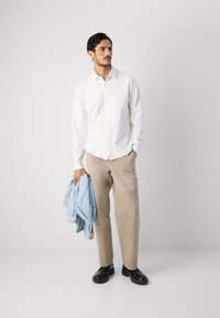 Witte blouse, beige broek, houdt een lichtblauwe spijkerjack vast, zwarte schoenen, staat op een neutrale achtergrond. Casual outfit.