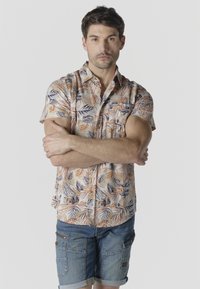 Hombre con los brazos cruzados, vistiendo una camisa de manga corta con estampado tropical y pantalones cortos de mezclilla azul, sobre un fondo claro y sencillo.