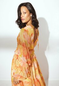 Frau mit schulterlangem dunklem Haar, die ein langes gelb-orange-blümchen Kleid mit Rüschensäumen trägt, seitlich vor einem weißen Hintergrund gedreht.