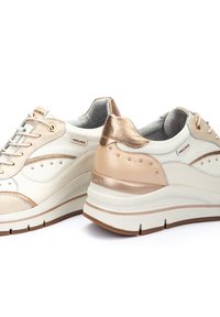 Zapatillas beige y crema con acentos metálicos, punta redonda, diseño con cordones, superposiciones laterales con patrón y suela gruesa blanca con base marrón.
