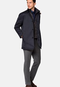 Navy blue outer coat with a button closure, paired with a dark quilted vest, gray trousers, and brown shoes. Smooth texture and classic design.