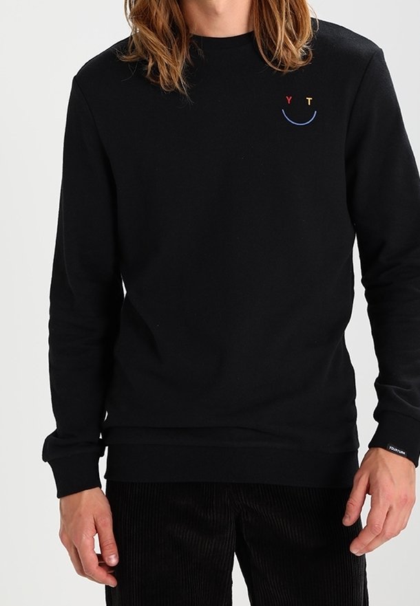 Svart långärmad tröja med liten broderad smiley-ansikte på vänster bröstficka, med röd "Y," gul "T," och blå böjd linje.