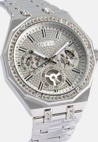 Silverfärgad oktagonal klocka med en kristallbeströdd bezel, texturerad urtavla, svarta timmarkörer och tre sub-dialer på ett metallarmband.