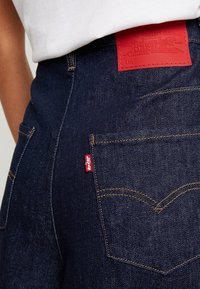 Tummansiniset farkut, joissa on punainen suorakaiteen muotoinen nahkalaatta vyötäröllä, pieni punainen Levi's-tunnus takataskussa ja kultaiset ompeleet.