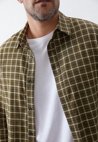 Groen-bruine geruite overhemd met knopen, gedragen over een wit T-shirt met ronde hals. Voorzien van een kraag en zwarte knopen. Geweven stof.