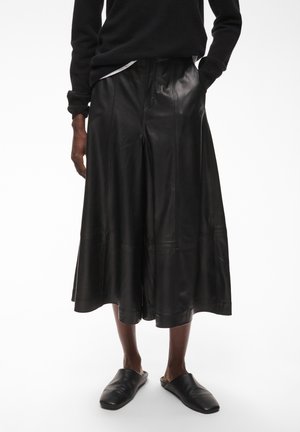 Personne portant un pantalon culotte en cuir noir à jambes larges, des chaussures noires sans lacets, un pull noir et une chemise blanche dépassant de la taille.
