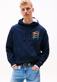 Tamsiai mėlynas džemperis su kišene, puošnus daugiaspalve "Tommy" logotipo siuvinėjimu. Dėvimas virš marškinėlių su apykakle ir šviesiai mėlynų džinsų.