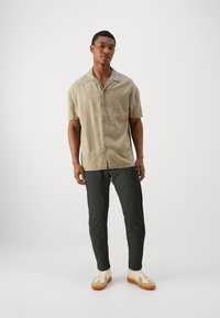 Camicia a maniche corte in lino color marrone chiaro con collo, chiusura a bottoni e vestibilità comoda, abbinata a pantaloni slim-fit verdi scuri e sneakers beige.