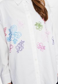 Cămașă albă cu nasturi, prezentând broderie florală colorată în roșu, albastru, mov, verde și roz. Fabricată dintr-un material neted și ușor.