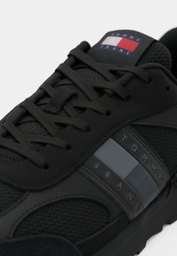 Black athletic shoe featuring mesh and synthetic materials, textured accents, tonal laces, and a logo patch in red, white, and blue.