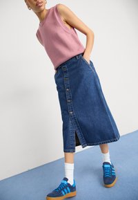 Fustă din denim cu nasturi în față, buzunare laterale și o fanta; asortată cu un top fără mâneci roz și adidași albaștri cu dungi în contrast.