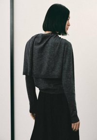 Femeie cu păr drept, negru, îmbrăcată într-un pulover stratificat de culoare gri închis și fustă neagră, stând în fața unui fundal deschis, orientată spre lateral.