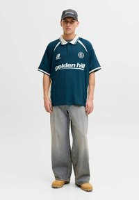 Jeune homme portant un polo de sport bleu marine avec le texte « golden hill », un jean ample clair, des bottes marron clair et une casquette noire sur un fond blanc uni.