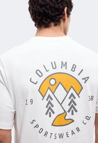 Άνδρας που φοράει λευκό T-shirt με κίτρινο και γκρι γραφικό βουνό, δέντρα, ήλιο και ποτάμι και κείμενο "Columbia Sportswear Co. 1938" στην πλάτη.