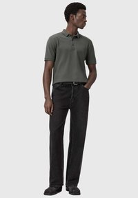 Uomo che indossa una polo a maniche corte verde scuro e jeans neri a gamba dritta, in piedi con una mano in tasca, guarda verso destra.