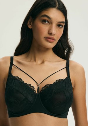 Kobieta o długich ciemnych włosach, ubrana w czarny koronkowy biustonosz z cienkimi ozdobnymi ramiączkami i małym złotym zapięciem w kształcie litery V na środku.