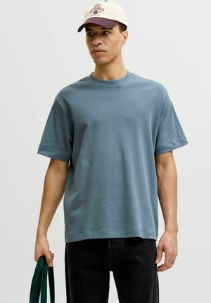 Homme portant un t-shirt surdimensionné bleu clair et une casquette de baseball beige tenant un sac fourre-tout vert sur fond blanc.