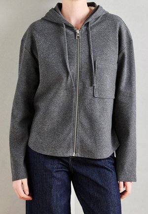 Persoon draagt een grijze zip-up hoodie met een borstzak en donkerblauwe spijkerbroek, staand tegen een effen lichte achtergrond.