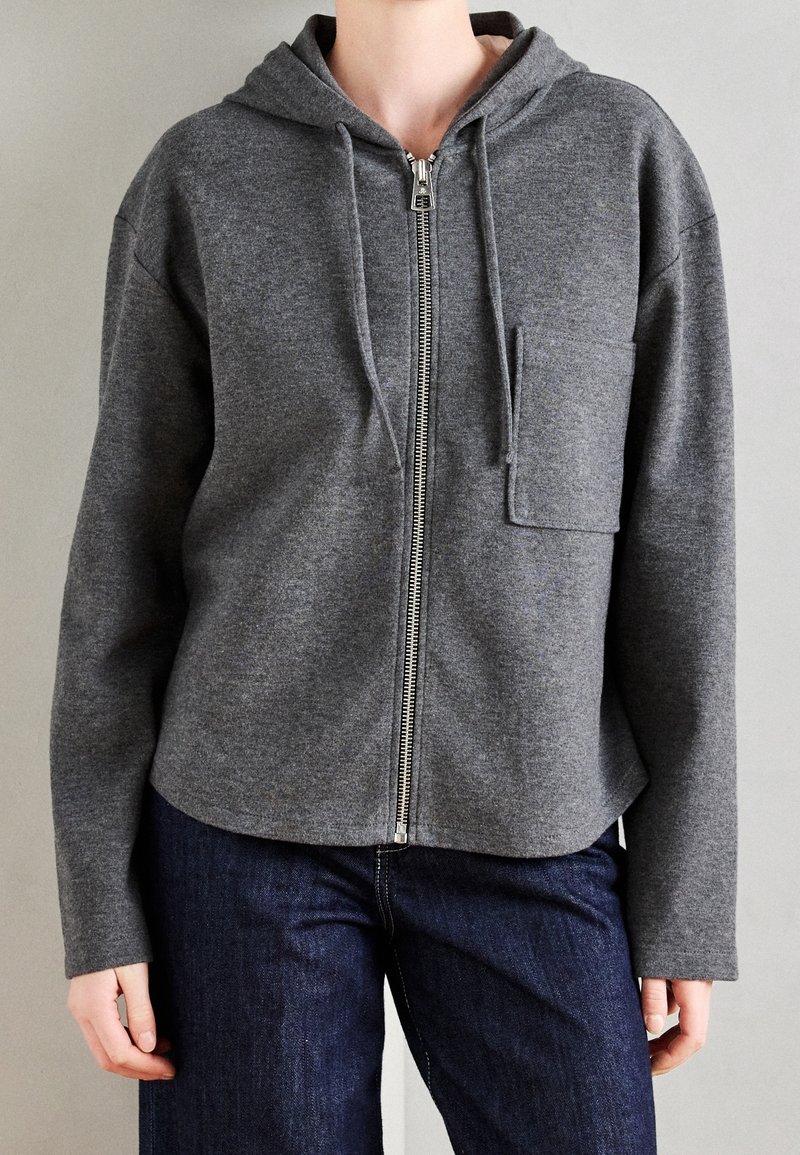 Personne portant un sweat à capuche gris zippé avec une poche poitrine et un jean bleu foncé, debout devant un fond clair uni.