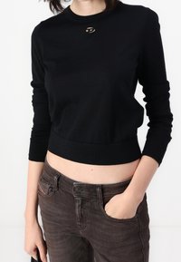 Czarny krótki sweter z okrągłym dekoltem i długimi rękawami, z małym dekoracyjnym metalicznym akcentem przy kołnierzu, w połączeniu z brązowymi dżinsami.