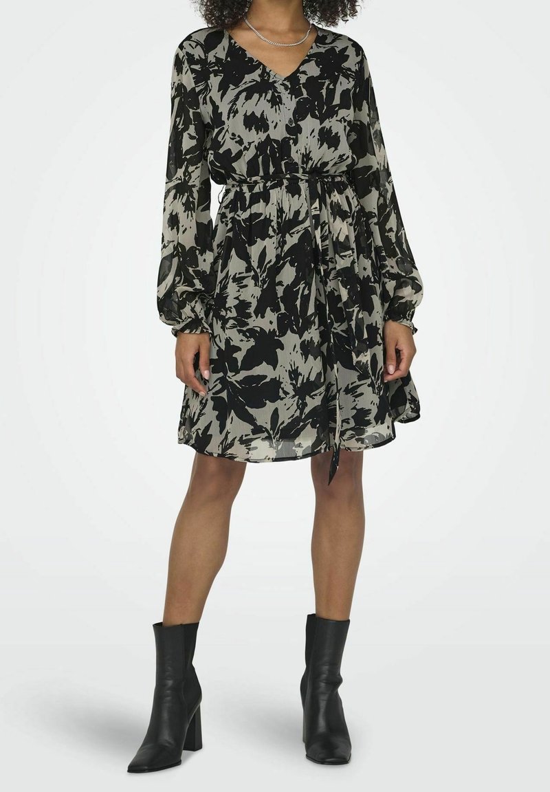 Femme portant une robe longue à manches longues noire et grise à motif floral, arrivant au genou, avec une ceinture à la taille et des bottines noires à talons.