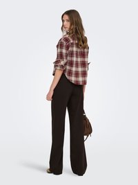 Jeune femme aux cheveux ondulés portant une chemise à carreaux retroussée, un pantalon noir à jambes larges, des chaussures beiges, tenant un sac à main marron, regardant par-dessus son épaule.