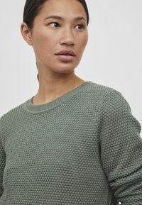Pull en maille verte texturée avec un col rond. Présente un motif léger en nid d'abeille et des poignets côtelés. Matière lisse et douce.