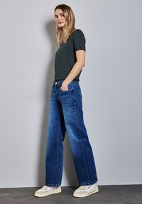 Tmavě zelená košile s krátkým rukávem v kombinaci s širokými modrými džíny. Tenisky s béžovým a bílým designem doplňují celkový vzhled.