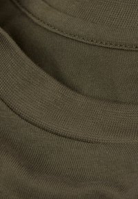 Olivengröna bomullstyg med en slät textur, som har flera kurviga u-ringningar och fina ribbade kanter.