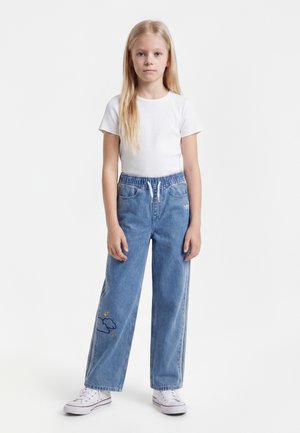Jeune fille aux longs cheveux blonds portant une chemise blanche à manches courtes, un jean bleu à jambes larges avec une broderie de nuages, et des baskets blanches, debout.