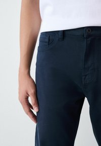 Pantaloni in misto cotone blu navy dal taglio slim, con tasca frontale, dettagli di cuciture sottili e chiusura con cerniera e bottone.