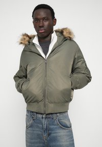 Oliivroheline pommikindel jakk, millel on faux karvapalaga kapuuts, lukuga sulgemine ja soonikkäised, seljas halli särk ja sinised teksad.