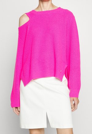 Nainen, joka pukeutuu kirkkaan vaaleanpunaiseen neulepaitaan, jossa on yksi hartia paljaana, ja valkoiseen polvipituiseen hameeseen, jossa on etuslitsi.