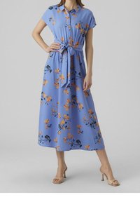 Vaaleansininen kukalline maxi-mekko, jossa on lyhyet hihat, vyötäröllä vyö ja oransseja kukkia. Sujuva kangas rentoa istuvuutta varten ja laskeutuva hame.