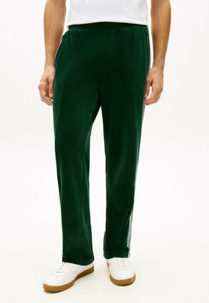 Mężczyzna ubrany w ciemnozielone aksamitne spodnie z bocznymi paskami, białe trampki i białą koszulę, stojący na tle jednolitego białego tła.