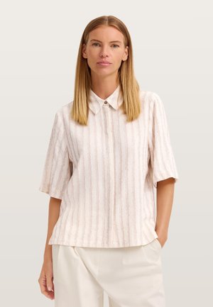Frau mit glatten blonden Haaren trägt eine kurzärmelige, hellbeige Bluse mit vertikalen Streifen und weiße Hosen, steht mit einer Hand in der Tasche.