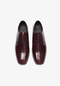 Zapatos de vestir de cuero burdeos con un acabado brillante, que presentan cordones negros y una puntera redondeada, sobre un fondo blanco.