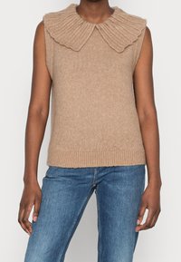 Maglione senza maniche beige con un ampio colletto a coste, dalla texture morbida e orlo aderente. Indossato con jeans di denim.