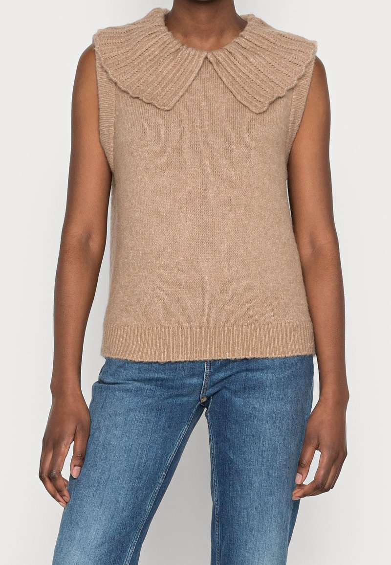 Maglione senza maniche beige con un ampio colletto a coste, dalla texture morbida e orlo aderente. Indossato con jeans di denim.