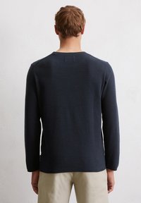 Granatowy sweter z długim rękawem o fakturowanym dzianinowym wzorze, z prążkowanym dekoltem i dołem, w zestawieniu z jasnobeżowymi spodniami w tle.