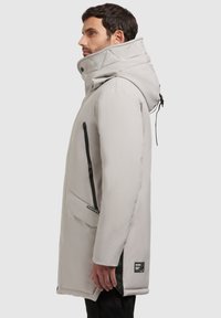 Parka impermeabil de culoare gri deschis, cu guler înalt, glugă reglabilă, buzunar lateral cu fermoar și accente texturate pe glugă.