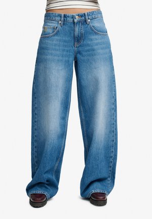 Jean en denim bleu à jambes larges porté par une personne avec des chaussures foncées, haut rayé court visible et légère décoloration au niveau de la cuisse.