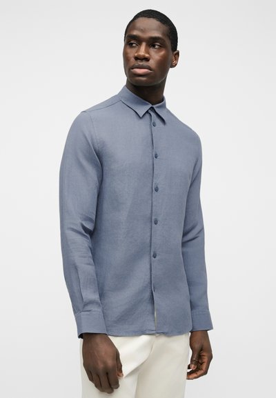 Άνδρας που φορά μπλε πουκάμισο με μακριά μανίκια και κουμπιά, και λευκό παντελόνι, κοιτάζοντας ελαφρώς προς τα αριστερά του, με φόντο μια απλή επιφάνεια.