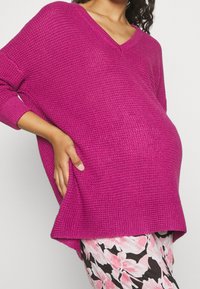 Gravid kvinna som bär en texturerad magenta tröja och blommönstrade byxor, håller sig om sidan med ena handen.