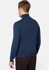 Granatowy sweter z golfem wykonany z miękkiego dzianiny, charakteryzujący się dopasowanym krojem i ściągaczami przy mankietach, noszony z brązowymi spodniami.