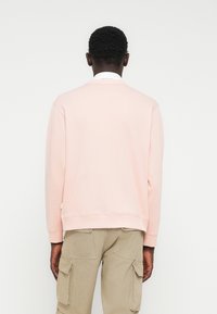 Hombre de piel oscura con sudadera rosa pálido y pantalones cargo beige, de pie frente a una pared blanca con las manos a los lados.