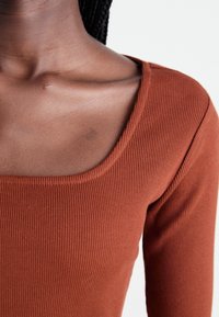 Close-up van een persoon met een roestkleurig geribbeld langarmshirt met een vierkante halslijn, waarbij de nek en het sleutelbeen zichtbaar zijn.