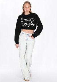 Junge Frau lächelt, trägt einen schwarzen, kurz geschnittenen Pullover mit dem Aufdruck "Smile everyday" und weiten, hellen Jeans. Sie geht mit einer Hand in der Tasche.