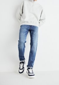 Sudadera con cremallera de color gris claro, jeans rectos de color azul y zapatillas negras y blancas con detalles de malla. Outfit simple y casual sobre un fondo neutro.