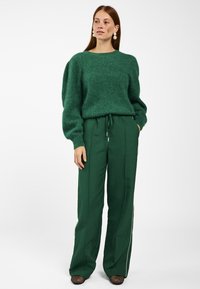 Vrouw met rood haar, die pareloorbellen draagt, een groene fuzzy trui, groene trekkoordbroek en zwarte netpantoffels, staand met haar handen in haar zakken.